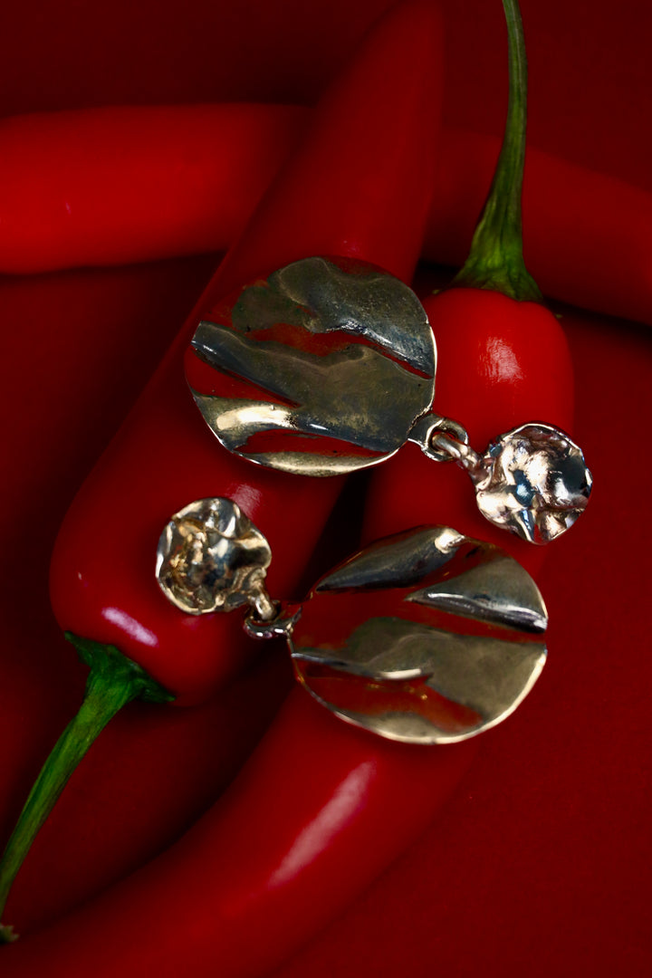 The Tide Drop Earrings by Madeleine Sculptural Jewellery—round and textured in silver—are shown atop red chili peppers against a matching background, their shiny, irregular surfaces reflecting light for a striking effect.