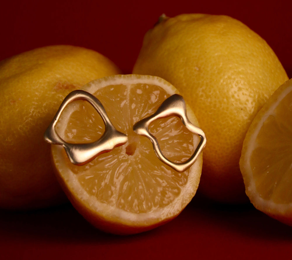 The Molten Studs by Madeleine Sculptural Jewellery—abstract gold earrings made from recycled brass—are displayed on a halved lemon with whole lemons in the background against a dark red backdrop.