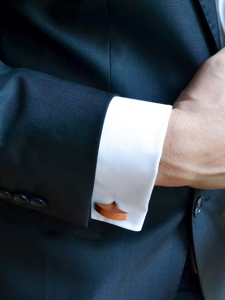 A close-up of a person wearing a dark suit jacket and white dress shirt. The cufflink, crafted from anodized aluminum and shaped like a mustache, is visible. The person's hand is partially in view, adjusting the suit jacket—an elegant nod to modern accessories with Filip Vanas's Secant Cufflinks.