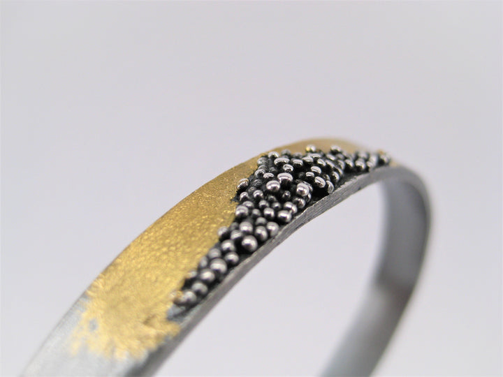 Close-up image of a metallic ring with a unique design. The Oxidised Etrusc Bracelet by MarCucurella features a smooth section of 24 kt yellow gold combined with an Etruscan granulation area consisting of multiple small oxidized silver spheres, creating an intricate and contrasting pattern. The background is blurred, focusing attention on the bracelet.