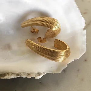 A pair of Inner Island Strata Large Earrings featuring textured gold ridged detailing rests on a white seashell dish, elegantly displayed on a marbled surface.
