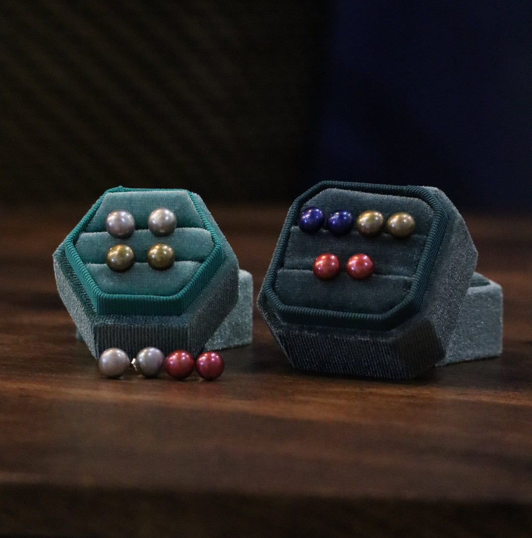 Two velvet ring boxes by The Collective Dublin showcase Pearl Earrings in gold, silver, purple, pink, and red on a dark wooden surface with a blurred background.