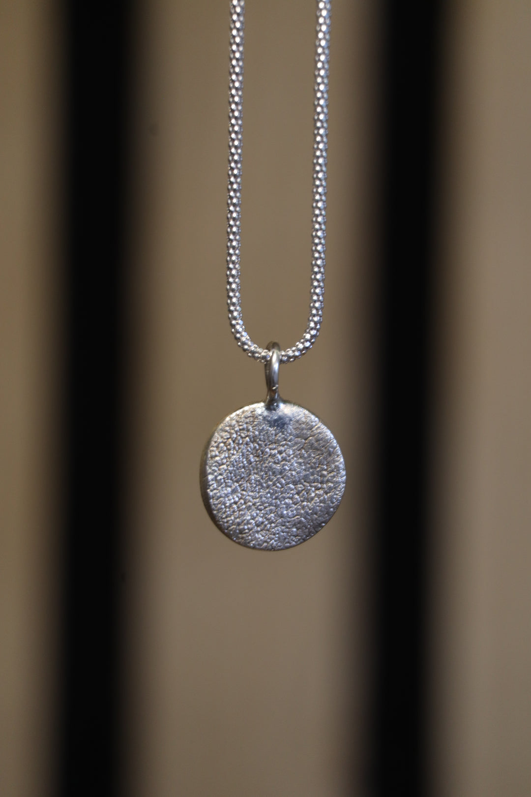 A close-up of the Silver Works Dog Nose Pendant Kit—a textured, round sterling silver pendant on a delicate chain—set against a neutral background with two vertical dark lines.