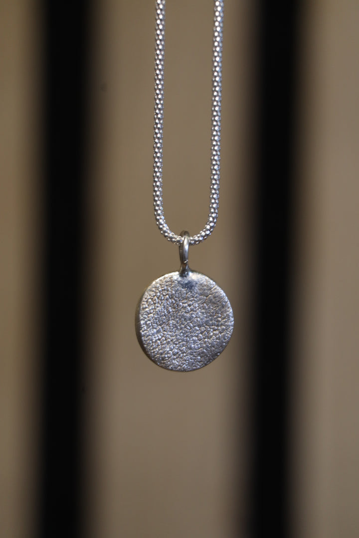 A close-up of the Silver Works Dog Nose Pendant Kit—a textured, round sterling silver pendant on a delicate chain—set against a neutral background with two vertical dark lines.