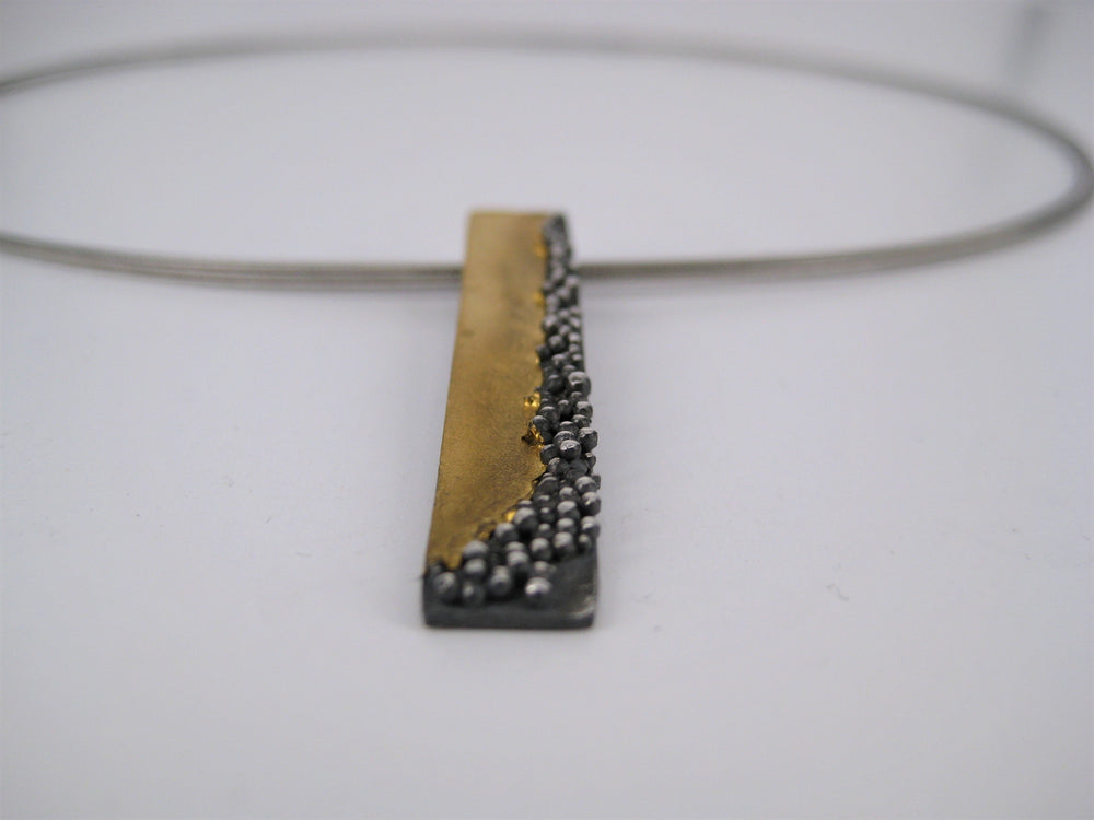 A close-up of a modern necklace featuring a rectangular pendant on a wire choker. The Etrusc Pendant by MarCucurella, adorned with 24 kt yellow gold on the left and oxidized silver with small metallic spheres reflecting Etruscan granulation on the right, stands out against the plain white background.