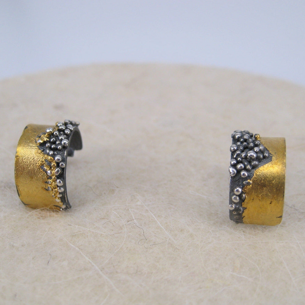 A close-up image of two gold and silver textured earrings set against a neutral background. The Etrusc Small Hoop Earrings by MarCucurella feature a rough, organic design with small bead-like details of oxidized silver clustered on the surface of the 24 kt yellow gold bands, reminiscent of Etruscan granulation.