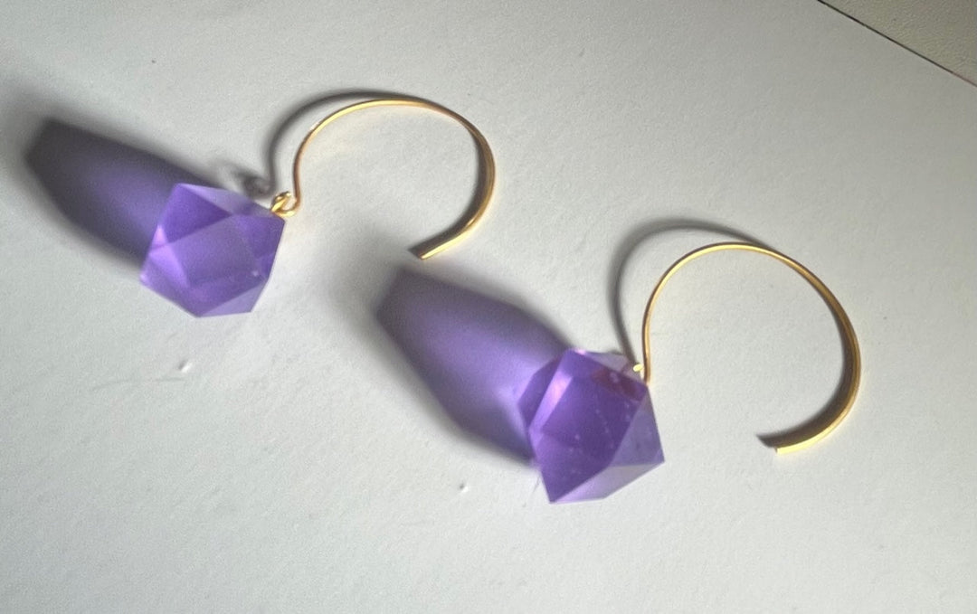 Beira by Irene Reyes' GEO Round Hook Earrings, featuring gold hooks and purple faceted gem-like ornaments, rest on a white surface. Their geometric design casts unique shadows, adding depth to the image.