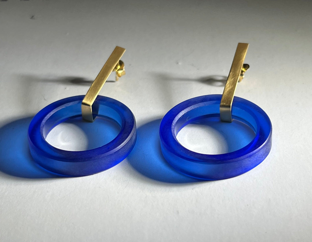 A pair of Soul Bottle Long Circle Earrings featuring blue circular rings made from recycled bottle glass attached to gold rectangular bars from Beira by Irene Reyes. The translucent blue rings cast soft shadows on the light-colored surface beneath them. The gold bars attach to 925 silver base earring posts for secure wear.