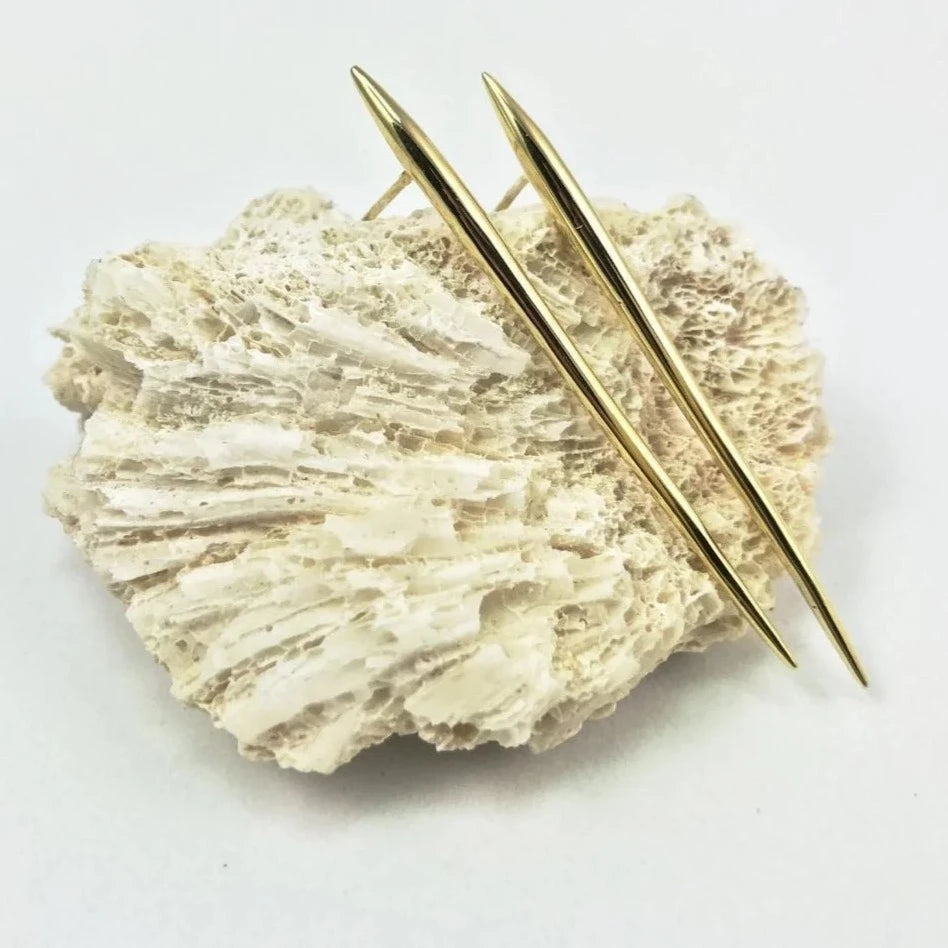 A textured off-white coral fragment is shown on a white background, with two Jennifer Kinnear Sea Urchin Spine Earrings - Gold in various sizes arranged diagonally across it.