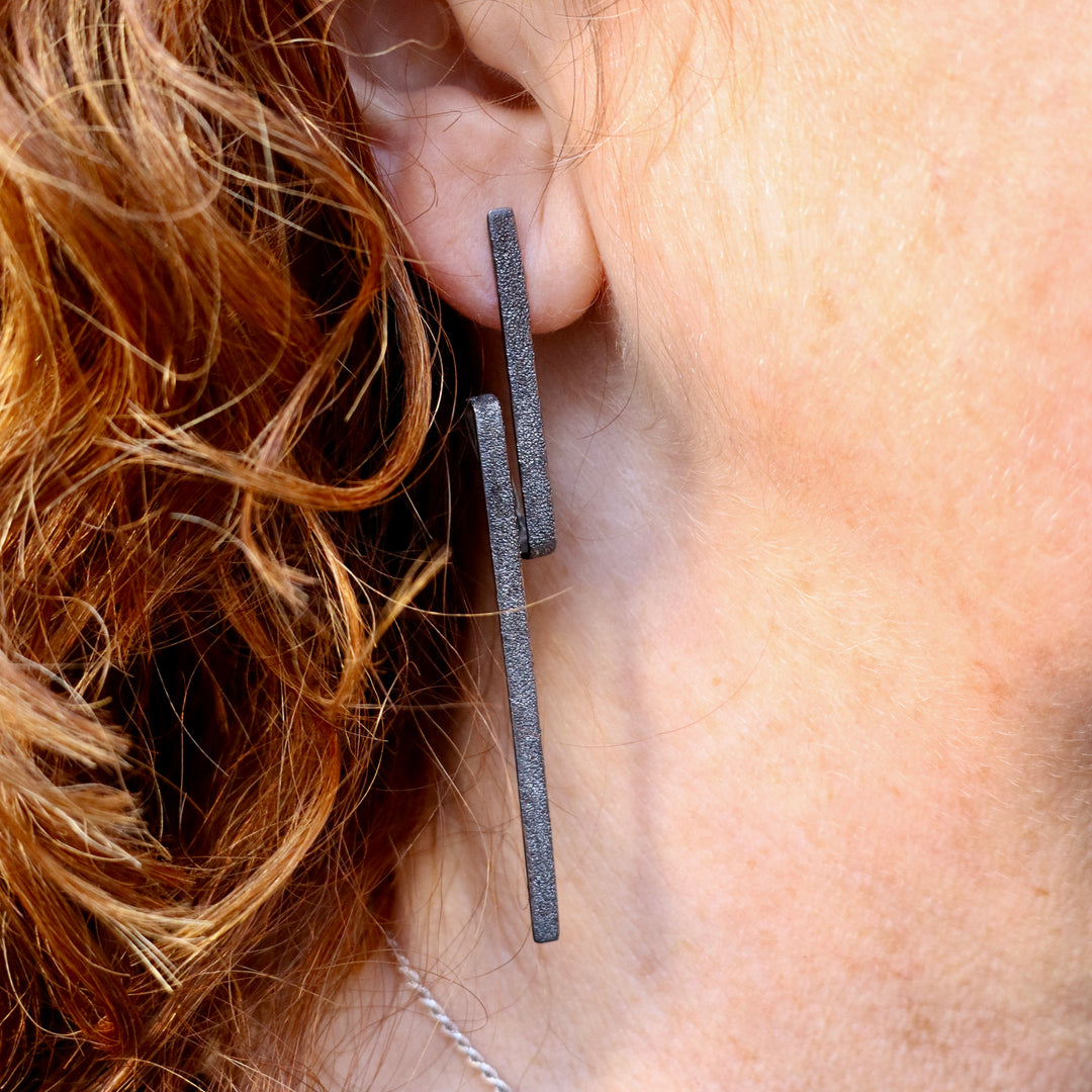 Close-up of a person with curly red hair wearing Theodora D Lysithea Black Rhodium Silver Earrings, featuring two matte black sterling silver rectangular bars in a modern, minimalist geometric design hanging vertically along the ear.