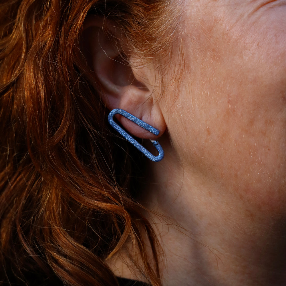 Close-up of a person with red hair wearing Theodora D's O Silver Earrings with Blue Ceramic Coating, handcrafted in Greece. The image highlights the unique jewelry and the texture of their skin and hair.