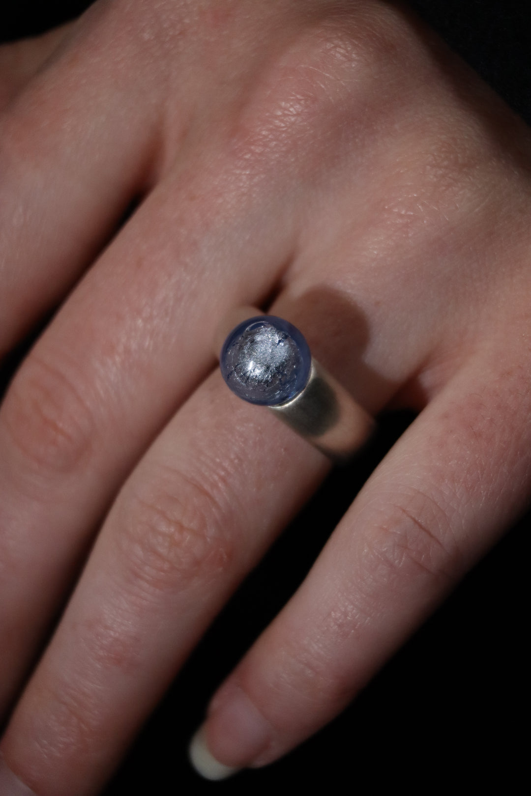 A close-up of a hand wearing the Detlef Menard Muranoperle Silver Ring 12mm, featuring a handcrafted sterling silver band with a round, translucent blue stone, set against a dark background.
