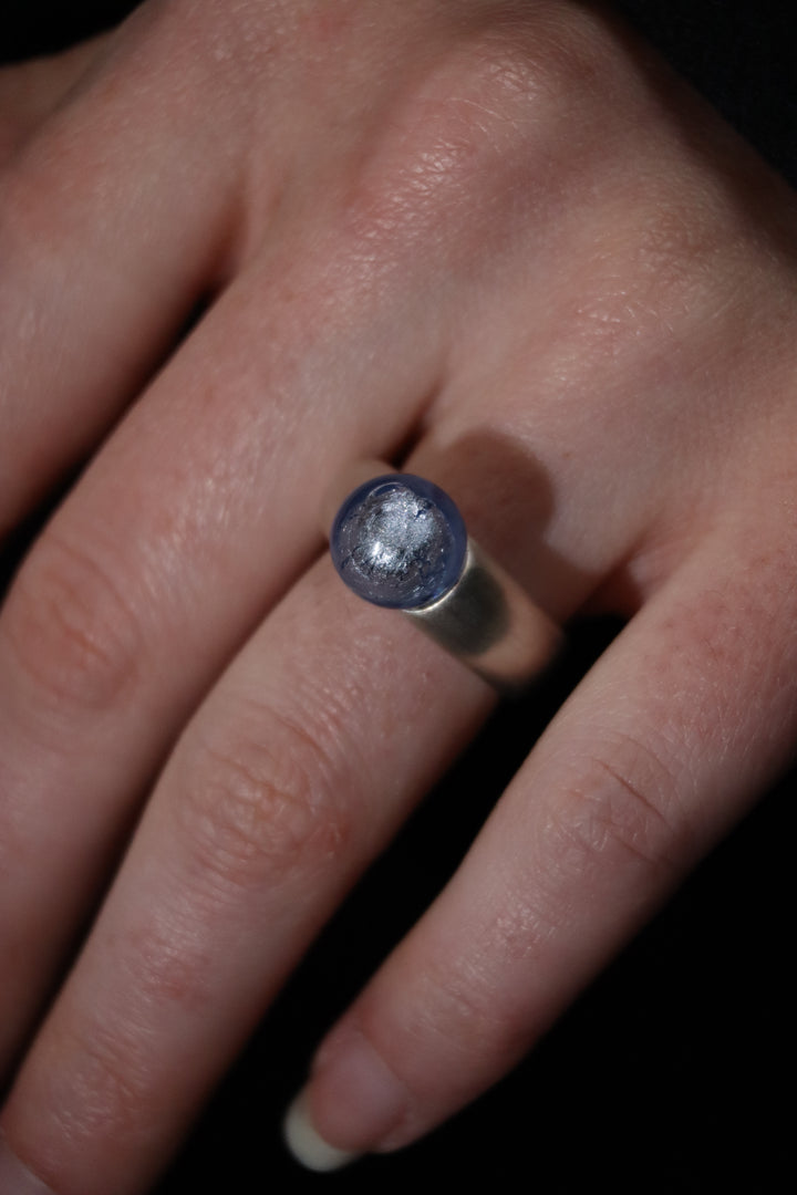 A close-up of a hand wearing the Detlef Menard Muranoperle Silver Ring 12mm, featuring a handcrafted sterling silver band with a round, translucent blue stone, set against a dark background.