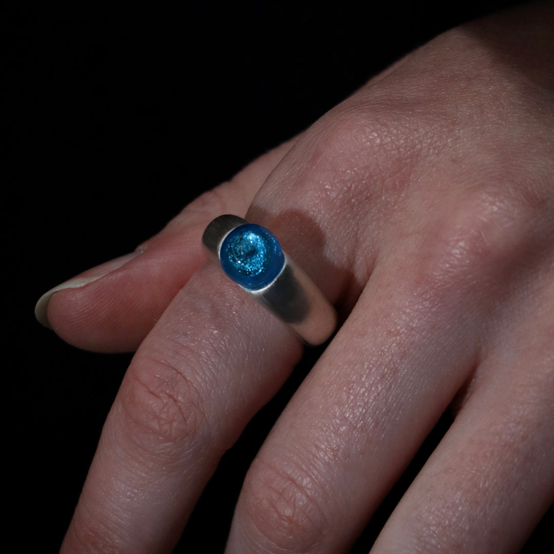 A close-up of a hand wearing the Detlef Menard Murano Half Perle Silver Ring 10mm, featuring a round, shiny blue Murano glass stone set in sterling silver, shown against a dark background.