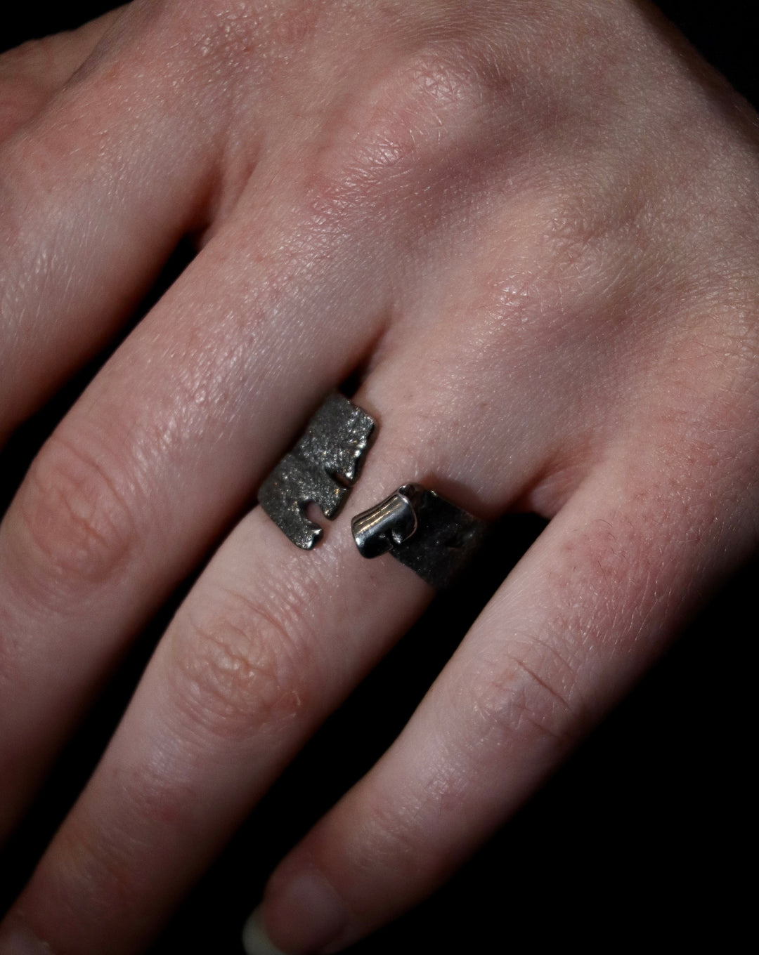 A close-up of a hand wearing Theodora D's Rythmos Black Rhodium Ring, featuring an open-ended design with rough, irregular edges, positioned across the fingers against a dark background.