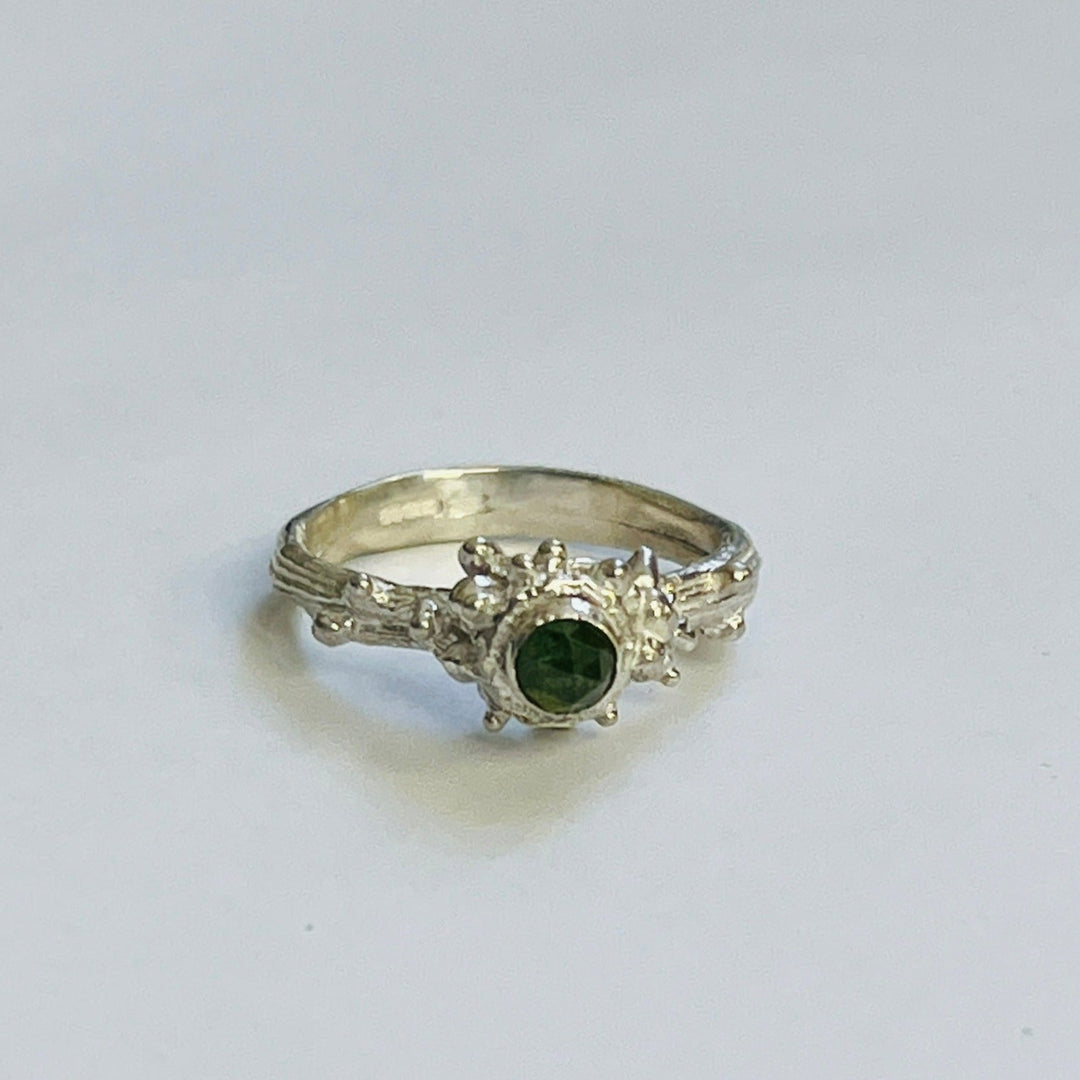 The Wild Woods Ring Green tourmaline by Abbie Dixon features a central green tourmaline gemstone flanked by small, clear stones on a sterling silver twig ring with intricate detailing on the band. The ring is displayed against a plain, light background.