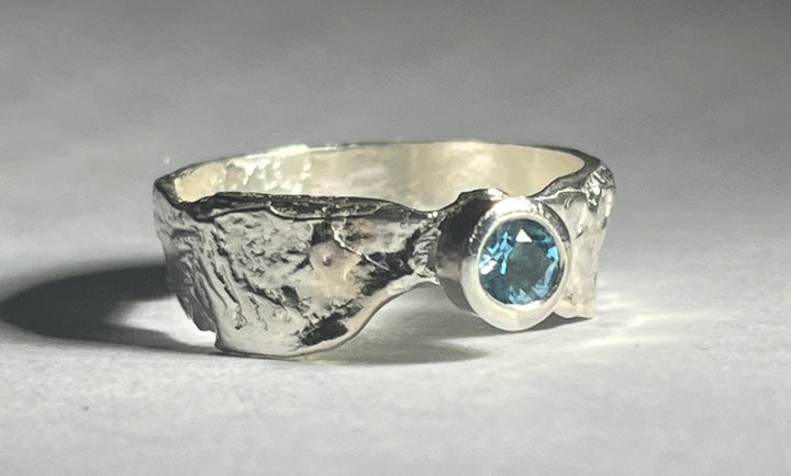 A textured silver Faerie Tale Ring Thin Band by Caroline Stokesberry-lee featuring an uneven, rugged design with a single blue topaz gemstone set in a round bezel. The ring reflects light in various shades, highlighting its intricate surface details and the vibrant blue of the gem.