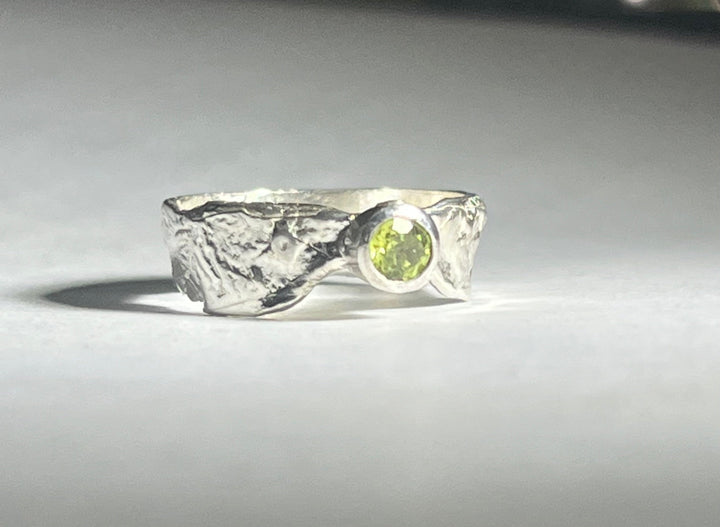 A silver Faerie Tale Ring Thin Band by Caroline Stokesberry-lee with a textured, rough surface features a small, round, bright green gemstone set in a bezel setting at the center. The ring sits on a smooth, light-colored surface with soft lighting casting shadows around it.