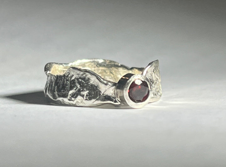 The Faerie Tale Ring Thin Band by Caroline Stokesberry-lee is a textured silver band with a rough, jagged surface design, featuring a small, round red gemstone set in a smooth bezel on one side. Positioned on a light, slightly reflective surface with a soft shadow cast behind it, this gem captivates with its mystical allure.