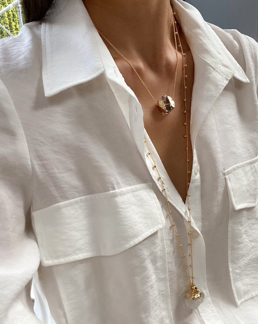 Close-up of a person wearing a white button-up shirt accessorized with layered 16k gold plated Aria V Khione Necklaces. The necklaces feature various pendant designs, including one with a textured, shiny gold sphere and another showcasing a clear quartz pendant. The shirt has pockets on both sides.