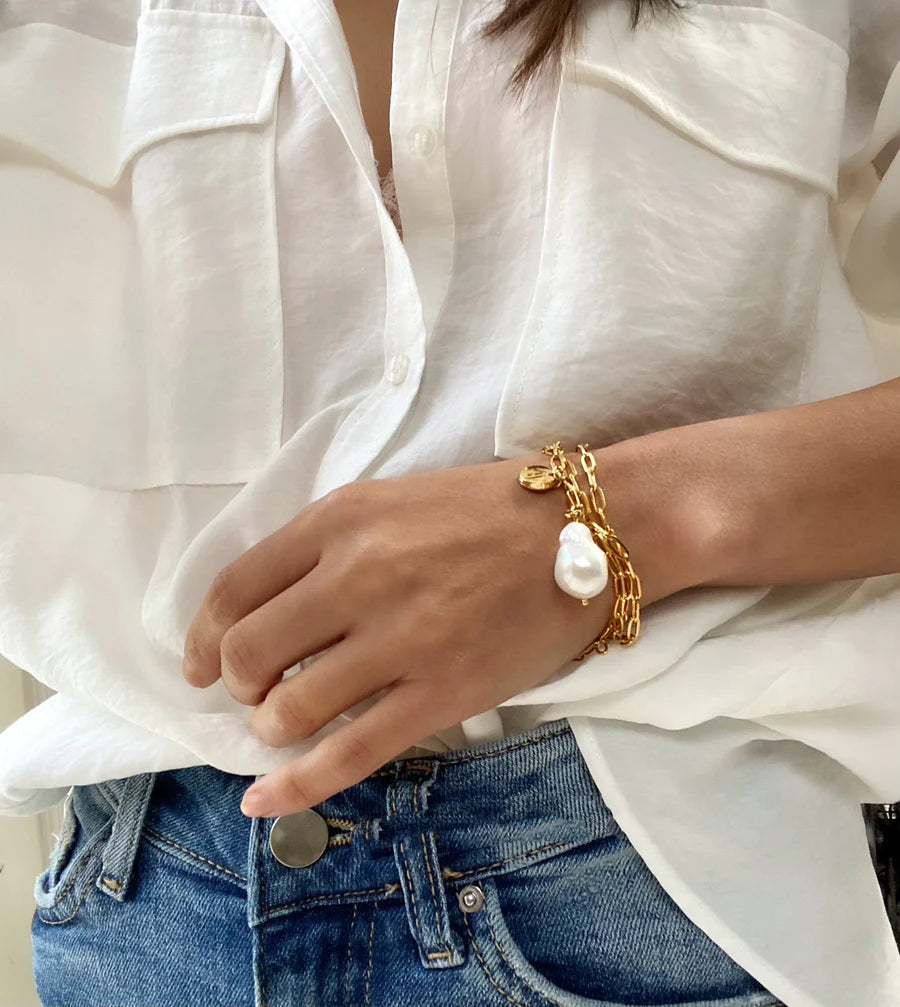 A person wearing a white button-up shirt and blue jeans has their hand resting on their hip, showcasing a Medea Necklace/Bracelet by Aria V adorned with a baroque white pearl and other charms.