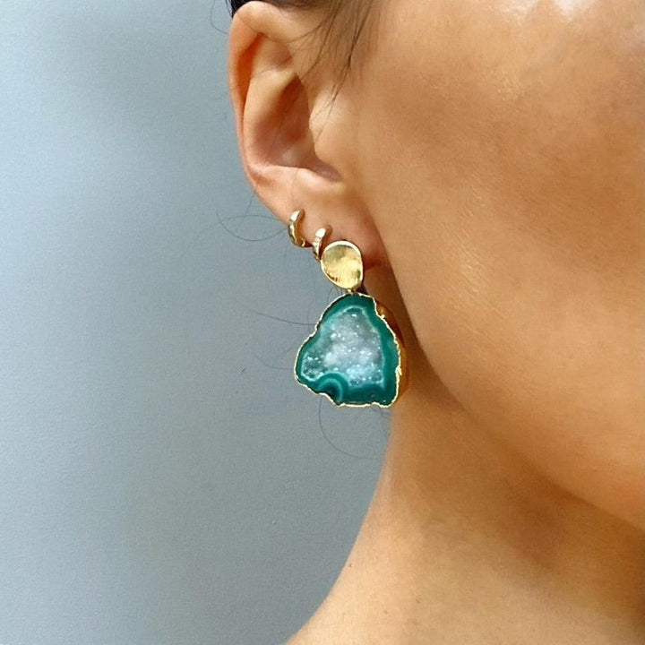 Close-up of a person's ear wearing a large, irregularly-shaped teal gemstone earring with a gold setting. The 24k gold plated Ileanna Earrings by Aria V consist of a circular stud at the top and a larger triangular-shaped gemstone below. Two small gold hoop earrings are also worn on the upper ear. Handmade in Ireland.