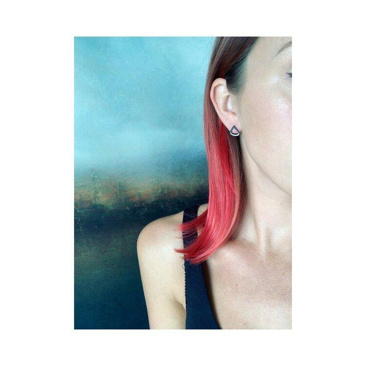 A person with shoulder-length red hair brushed back is shown from the neck up, slightly turned to the side. They are wearing a sleeveless black top and a triangular oxidised Leoma Drew Two Tone Silver Wing Stud Earrings. The background features an abstract gradient of blue and green hues.