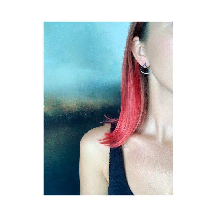 A person with straight, shoulder-length, pink-dyed hair brushed back is seen from the side, with their face partially out of frame. They are wearing a black sleeveless top and a single Two Tone Silver Wing Stud Earring by Leoma Drew. The background is an abstract blend of blue and brown colors.