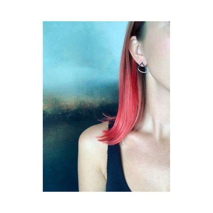 A person with straight, shoulder-length, pink-dyed hair brushed back is seen from the side, with their face partially out of frame. They are wearing a black sleeveless top and a single Two Tone Silver Wing Stud Earring by Leoma Drew. The background is an abstract blend of blue and brown colors.