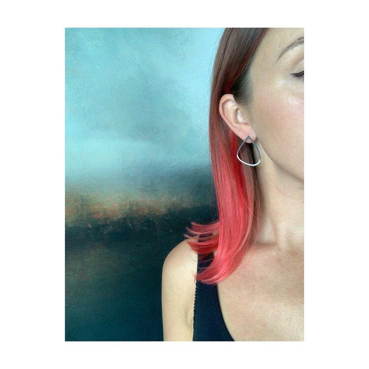 A person with pink ombre hair, brushed back stylishly, is wearing a black tank top and a Two Tone Silver Wing Stud Earrings by Leoma Drew. Only the right side of their face and hair are visible against a blurred, abstract background with blue and dark tones.