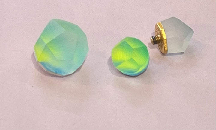 A pair of round, iridescent green and blue Fruit Bijoux Ring Top Aura earrings on a light pink surface. The earring on the right has a gold-tone backing attached while the left earring and its backing are separated. The faceted surfaces reflect light, showcasing vibrant colors, complemented by elegant sterling silver accents.