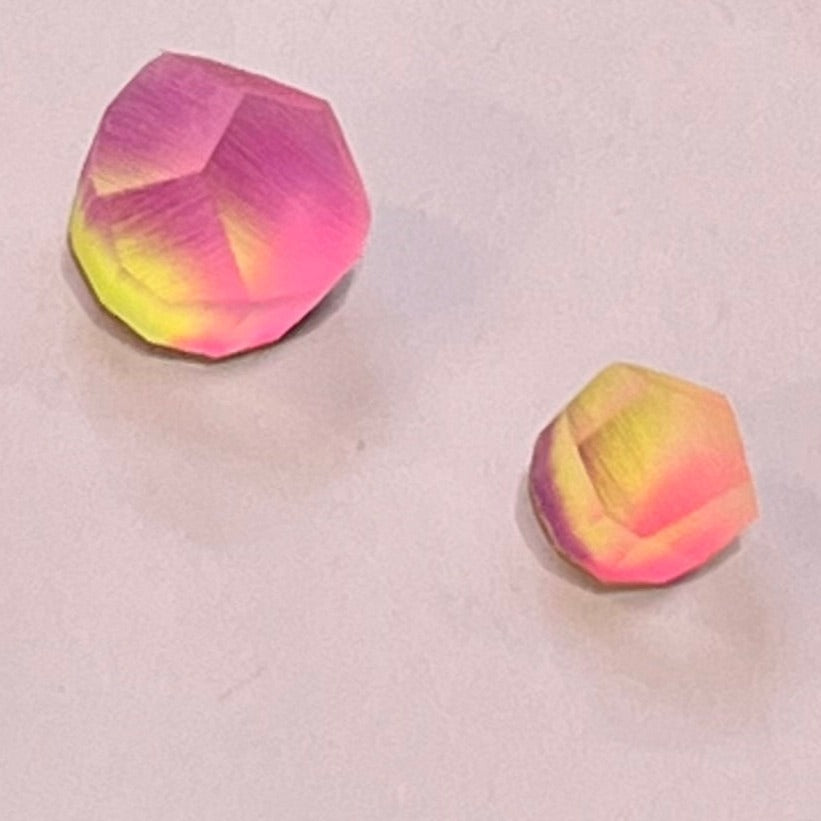 Two polyhedral objects with gradient colors ranging from pink to yellow on a light gray background. The object on the left, reminiscent of a sterling silver gem, is slightly larger than the one on the right. The colors blend seamlessly, giving them a vibrant and iridescent appearance. These objects are the Fruit Bijoux Ring Top Aura by Fruit Bijoux.