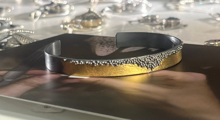 A MarCucurella Oxidised Etrusc Bracelet with a textured 24 kt yellow gold section sits on a reflective surface, surrounded by blurry background elements that include other jewelry pieces. The bracelet's design features a rough transition reminiscent of Etruscan granulation between the black and gold areas.