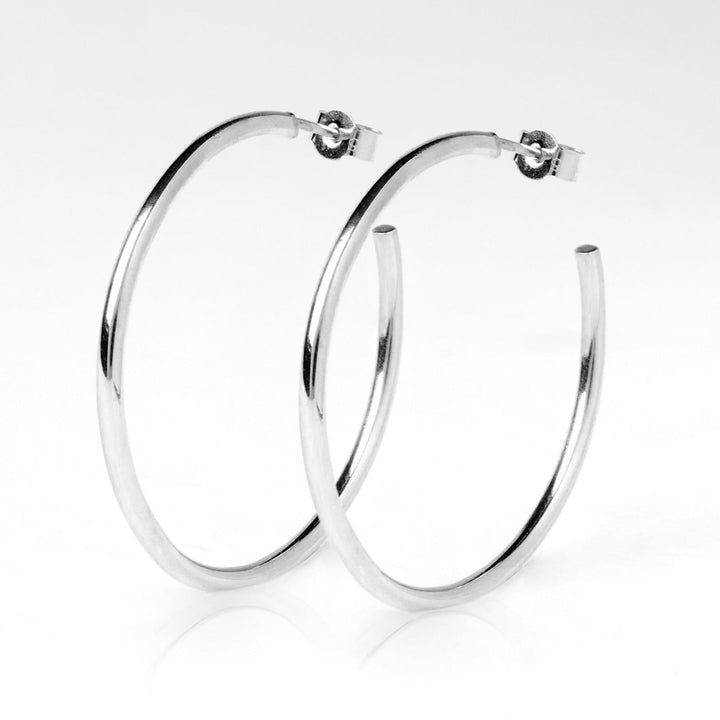 A pair of simple, elegant Cosmic Thread Hoops from Cosmic Boulevard, featuring a smooth and shiny silver surface. These circular earrings are designed with a small hinge and clasp mechanism at the top for easy securing. They are showcased against a plain white background.