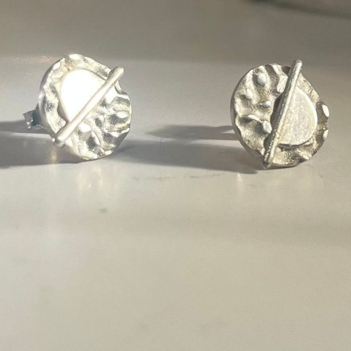 A pair of Lunar Eclipse Stud Earrings from Gallardo & Blaine Designs on a light surface. Each earring features an irregularly textured round design with an angled bar crossing the circle, creating a modern, abstract appearance reminiscent of Art Deco. The earrings are reflecting light, highlighting their shiny finish.