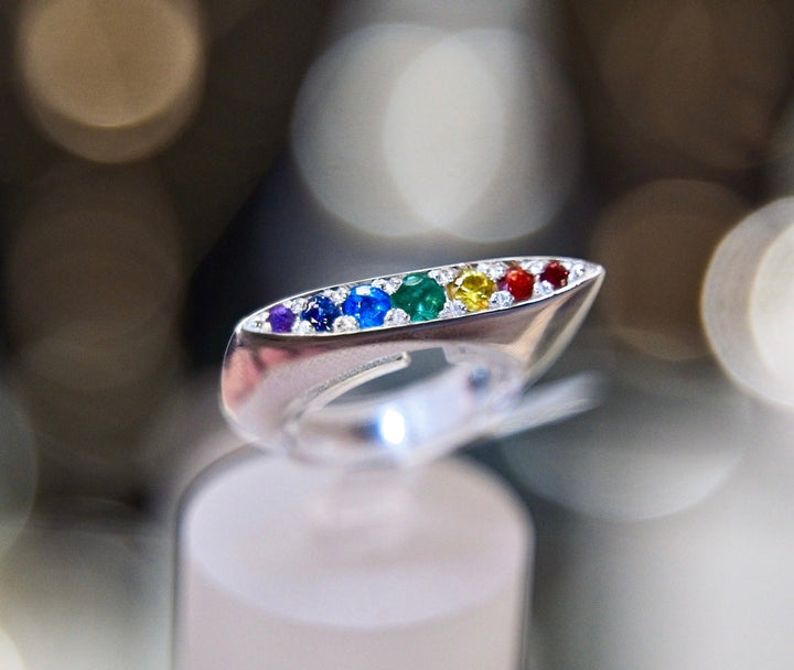 A close-up of a sterling silver Rainbow Star Ring by Cosmic Boulevard featuring an open oval design adorned with pavé set, multicolored gemstones. The stones are red, yellow, green, blue, and purple. An emerald stands out among them. The background is blurred with bokeh lights, creating a soft, dreamy effect.