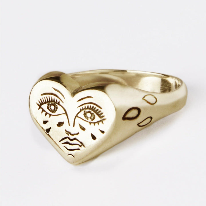 The Sad Lil Grimm Signet from Cosmic Boulevard is a handmade gold ring from Dublin, featuring a heart-shaped face with hand-engraved eyes, nose, and lips. Teardrops etched below the eyes give it a crying heart tattoo look, while the band boasts an uneven, organic texture.