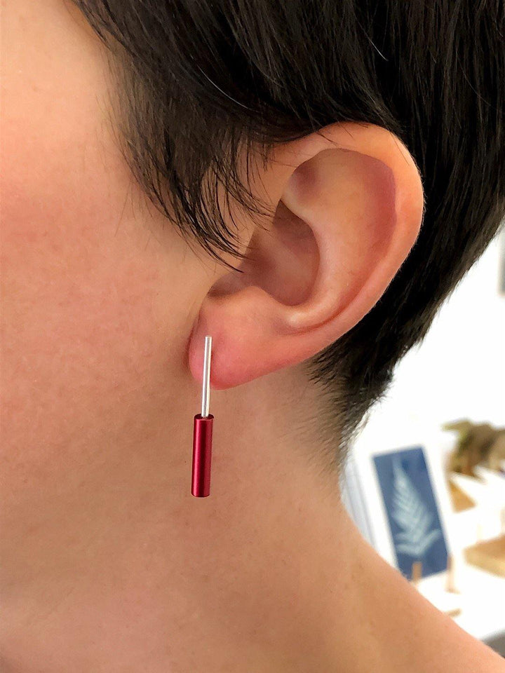 Close-up of a person wearing Filip Vanas Cylinder Drop Stud Earrings in sterling silver. The earring features a thin silver post with a small red cylindrical accent hanging from it. The person's short, dark hair partially covers the top of the ear, and the background is blurred.