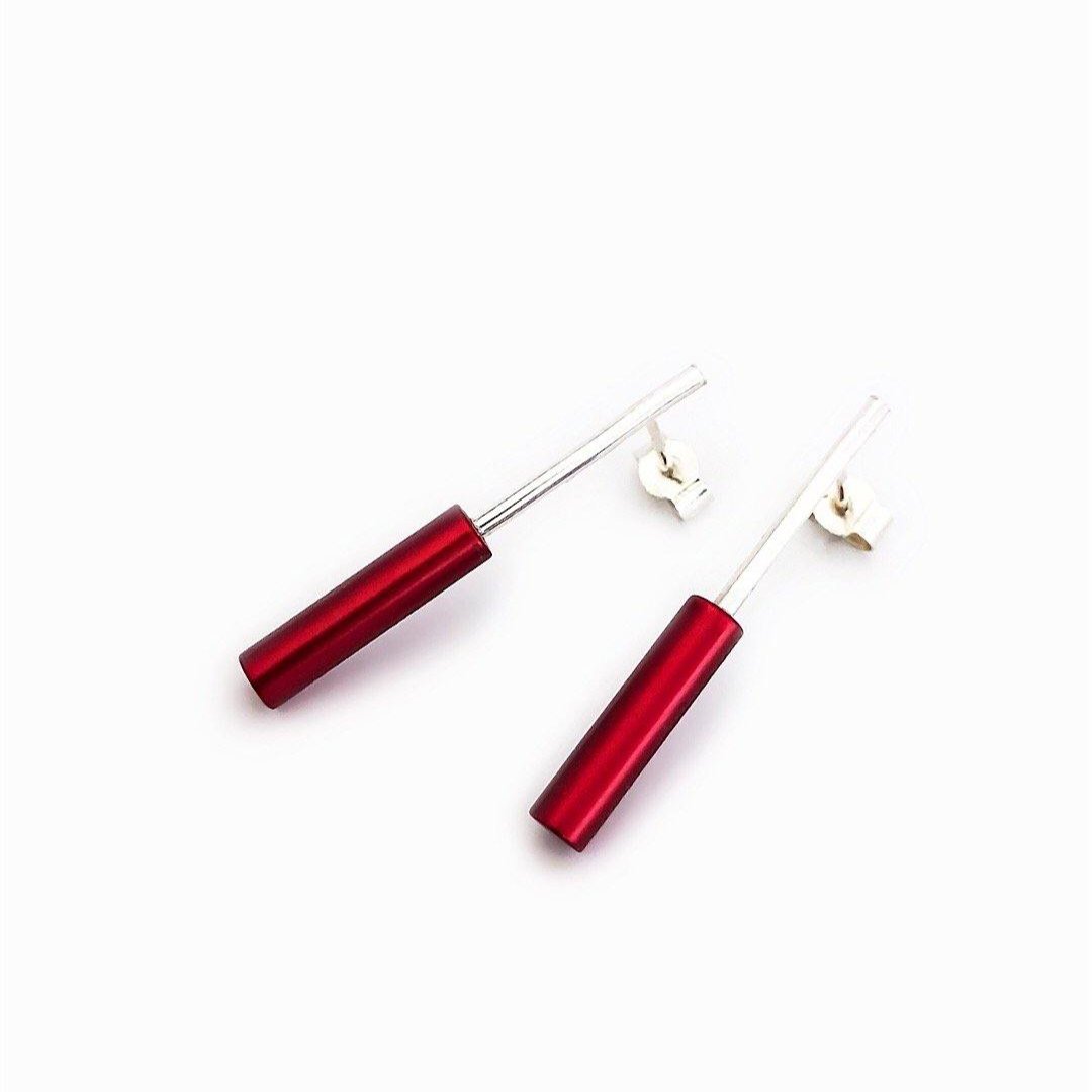 A pair of Cylinder Drop Stud Earrings in a sleek, modern design by Filip Vanas, featuring long, slender sterling silver posts with sophisticated red anodised aluminium cylindrical accents. The earrings come with white earring backs and are displayed against a plain white background.