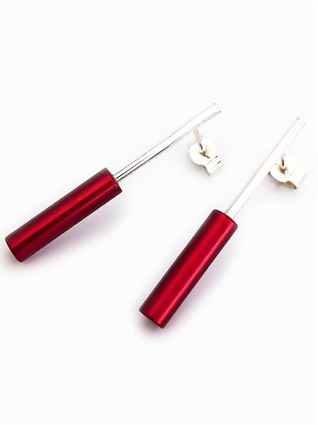 The Cylinder Drop Stud Earrings by Filip Vanas are a pair of modern, minimalist earrings featuring sleek, anodised aluminium red cylindrical pendants that hang from thin, sterling silver vertical bars. These stud fitting earrings are set against a white background and come with a simple clasp mechanism for fastening.