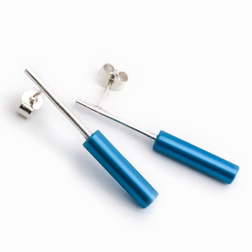 A pair of Cylinder Drop Stud Earrings by Filip Vanas, showcasing a modern and minimalist design with long, slender rods featuring cylindrical, matte blue ends. Each sterling silver earring includes a butterfly back closure for a sleek and contemporary look. The anodised aluminium earrings are elegantly displayed on a white background.