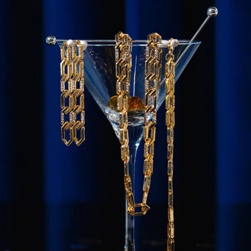 A martini glass on a dark surface, with four Pave & Metal Chain Link Earrings - Gold by Orelia London draped elegantly over the rim, showcasing their interlocking oval shaped links. The background features a rich blue curtain, creating a luxurious and sophisticated ambiance.