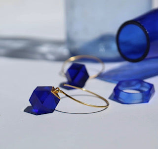 GEO Round Hook Earrings by Beira, featuring gold hoops and deep blue geometric pendants, rest elegantly on a white surface. In the background, matching blue shapes complement a Bullseye glass bottle, casting intriguing shadows.
