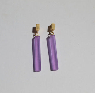 A pair of Soulbottle Straight Recycled Glass Earrings by Beira by Irene Reyes featuring long, translucent, purple bars made from recycled bottle glass, hanging from small, gold-colored rectangular studs on a plain white background.