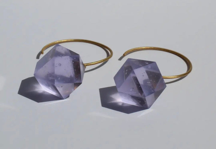 Beira by Irene Reyes offers GEO Round Hook Earrings, featuring faceted translucent purple stones resembling Bullseye glass on thin gold hoops, casting soft shadows on a light gray surface.