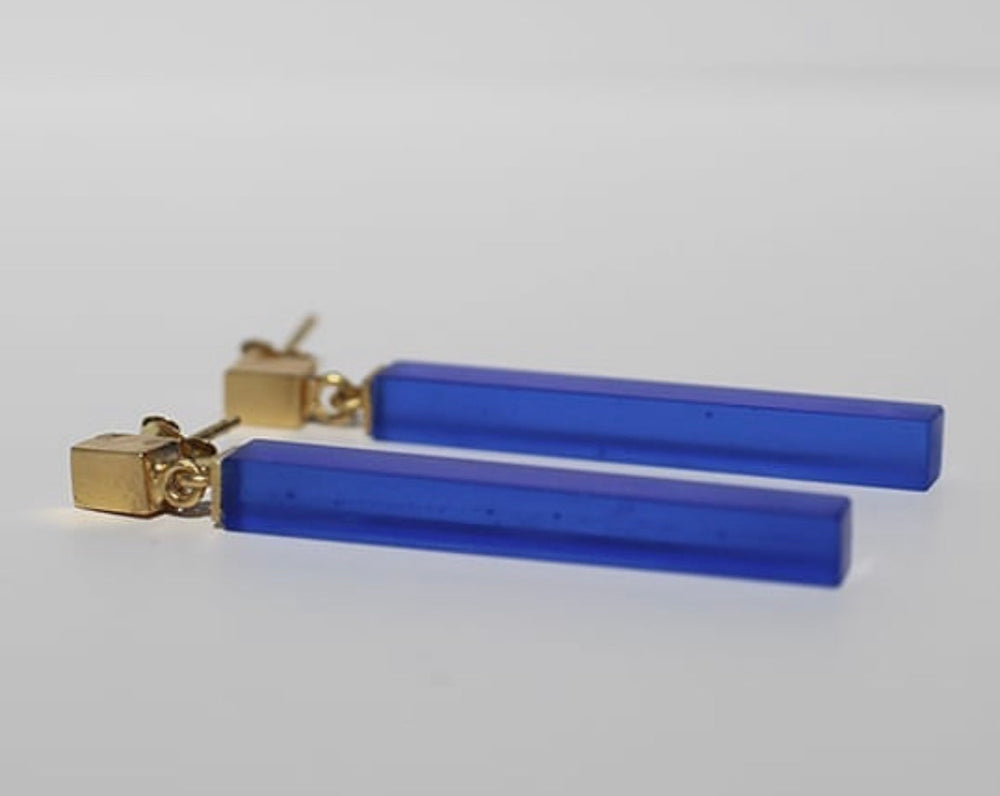 A pair of **Soulbottle Straight Recycled Glass Earrings** by **Beira by Irene Reyes** featuring long, rectangular blue pendants made from recycled bottle glass attached to small, gold square studs. The earrings are laid side by side on a plain white background.