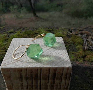 The GEO Round Hook Earrings are displayed with golden hoops and hexagonal green gems resting on a wooden box, against a soft, natural outdoor background of grass and trees. These earrings are from Beira by Irene Reyes.