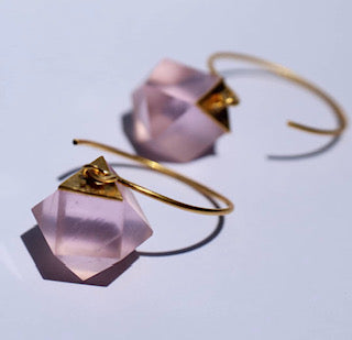 Gold wire earrings with matte, pink, faceted cube stones in a geometric design. Stones are linked by a gold square to the wire, casting soft shadows on a light surface. Ideal for fans of Beira by Irene Reyes's GEO Round Hook Earrings or unique Bullseye glass styles.