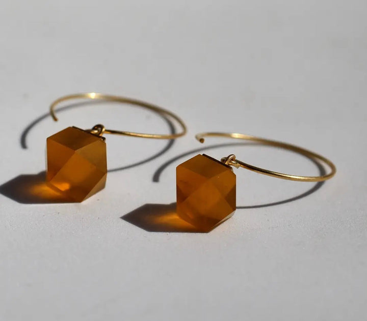 Beira by Irene Reyes' GEO Round Hook Earrings feature gold hoops with hexagonal amber-colored gemstones. These faceted gems hang elegantly, casting soft shadows on a plain, light surface and adding a hint of geometric charm.