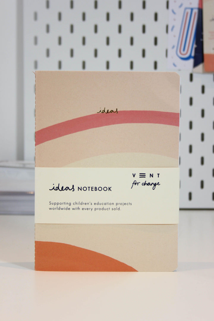 A beige notebook with wavy red and orange lines and the word "ideas" printed on the cover. The label reads: "Recycled Soft Cover A5 Notebook – Ideas Cream - Supporting children’s education projects worldwide with every product sold." Made from FSC certified paper, the brand "Vent For Change" is also displayed.